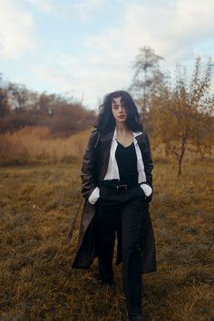 a woman standing in the middle of a field wearing black pants and a trench coat