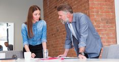 two people standing around a table looking at something on the paper that is in front of them