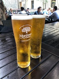 two glasses of beer sitting on top of a wooden table next to eachother