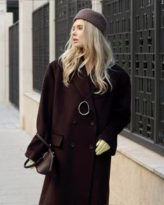 a woman in a black coat and hat is standing on the sidewalk with her hands in her pockets