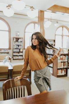 a woman standing in front of a table with a wooden chair and bookshelf behind her