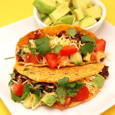 two tacos on a white plate with avocado and tomatoes in the background