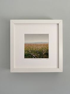 an empty white frame hanging on the wall above a field of grass and wildflowers