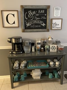 a coffee bar with cups and mugs sitting on it's side table next to a chalkboard