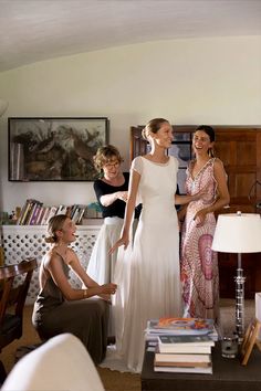 three women standing in a living room talking to each other and one woman wearing a white dress