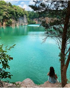 a woman sitting on top of a rock next to a tree near a lake filled with green water