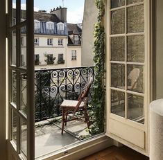 an open window with a chair on the balcony next to it and some buildings in the background