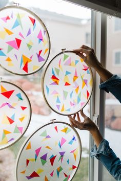 a woman is holding up some colorful paper art on a window sill and looking out the window