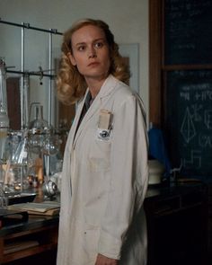 a woman wearing a lab coat standing in front of a blackboard