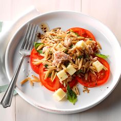 a white plate topped with pasta salad and sliced tomatoes next to a fork on top of a wooden table