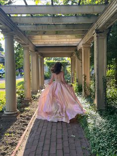 a woman in a long pink dress walking down a brick path under an arbor covered walkway