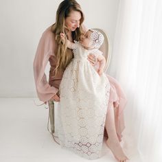 A woman in a flowy pink dress is sitting on a chair, holding a baby dressed in a long, white Poppy Christening Gown & Bonnet. They are both smiling and looking at each other. The background is bright and minimalistic, with sheer white curtains adding a soft touch. Cream Princess Gown Fitted, Cream Baptism Gown With Fitted Bodice, Princess Style Cream Wedding Gown, Cream Princess Wedding Gown, Princess Style Fitted Gown For First Communion, Princess Gown With Fitted Bodice For Baptism, Fitted Cream Gown For Pageants, Fitted Cream Gown For Pageant, Cream Gown With Lace Bodice For Pageants