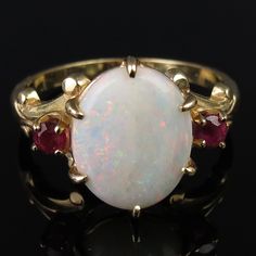 Pretty raspberry red rubies flank a natural white 1.68 carat Australian opal on this vintage 14k yellow gold ring. At the center is an oval 12mm x 9.8mm x 2.7 opal cabochon of white body and an array of color flashes. Fiery sparkles of greens, blues, reds and yellows come through when the gem refracts the light. The camera did not quite capture the actual beauty of the stone. The faceted 3mm pinkish-red rubies have an estimated total weight of .20 carat. The opal is held tightly with six sturdy Large Opal Ring, Oval White Ruby Gemstone Ring, Classic White Oval Ruby Ring, White Oval Ruby Ring, Oval White Ruby Ring, Oval Ruby Ring, Ruby Jewelry, Australian Opal, Opal Rings