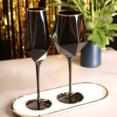 two black wine glasses sitting on top of a white plate next to a potted plant