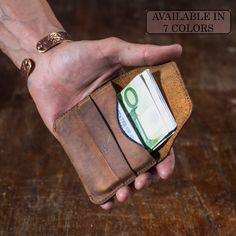 a hand holding a wallet with money in it on a wooden table next to a sign that says available in 7 colors