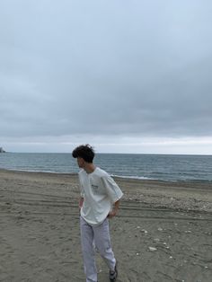 a young man standing on top of a sandy beach next to the ocean under a cloudy sky