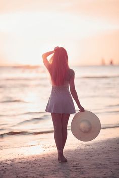 a woman walking on the beach at sunset with her hat over her head and hair blowing in the wind