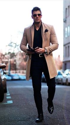 a man walking down the street wearing a tan coat and black pants with his hands in his pockets