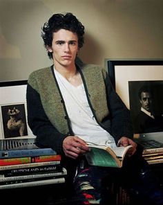 a young man sitting in front of a pile of books and holding an open book