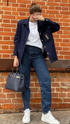 a woman standing in front of a brick wall holding a handbag and covering her face