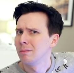 a man with black hair wearing a gray shirt and looking at the camera while sitting in front of a bed