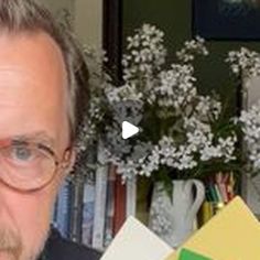 a man with glasses is holding a book in front of a flower vase and bookshelf