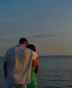 a man and woman standing on the beach looking out at the ocean with a kite flying in the sky