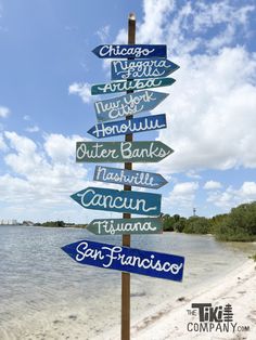 a wooden sign pointing in different directions on the beach with water and clouds behind it