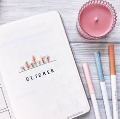 an open notebook sitting on top of a wooden table next to pens and a candle