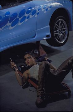 a woman laying on the ground next to a blue car and looking at her cell phone