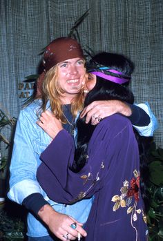 two people are hugging each other in front of a wall with plants and curtains behind them