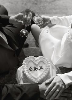 a man and woman sitting next to each other holding wine glasses in front of a heart shaped cake