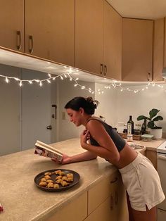 a woman is reading a magazine while standing at the kitchen counter with cookies in front of her