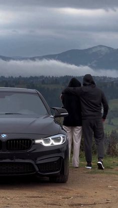 two people standing next to a black car on top of a dirt road with mountains in the background