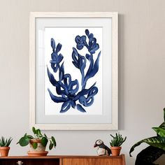 a blue plant in a white frame hanging on a wall next to potted plants