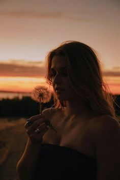 a woman holding a dandelion in her hand and looking at the sun setting