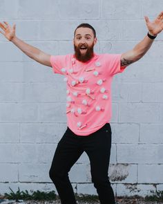 a man with his arms wide open standing in front of a white brick wall wearing a pink shirt and black pants