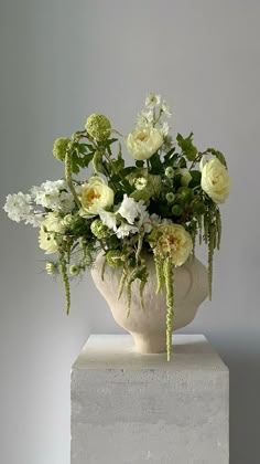 a white vase filled with lots of flowers on top of a block of concrete in front of a gray wall