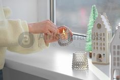 a person holding a lit candle in front of a window sill with miniature houses on the windowsill