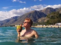 a man in the water holding a coconut drink and looking at the camera with mountains in the background