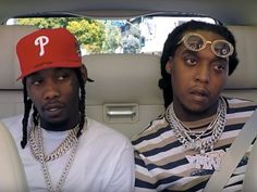 two young men sitting in the back seat of a car, one wearing a red hat