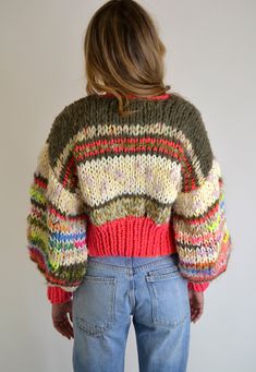 a woman standing in front of a white wall wearing a multicolored sweater and jeans