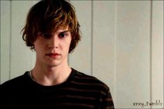 a young man standing in front of a white wall wearing a black and brown striped shirt