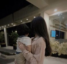 a woman holding a child in front of a building at night with lights on the windows