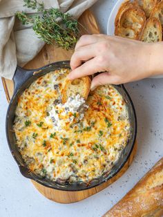 a person dipping cheese into a casserole dish with bread and herbs on the side