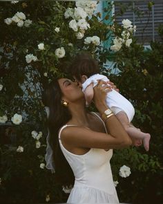 a woman in a white dress holding a baby up to her face with flowers behind her