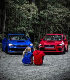 two people are sitting on the ground next to their cars in front of some trees