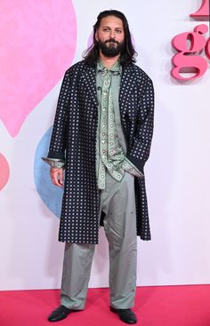 a man with long hair and beard standing in front of a pink heart background wearing an overcoat