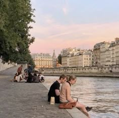 two people sitting on the edge of a body of water near some buildings and trees