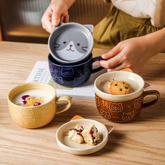 a person is holding a cat mug in front of two cups with food on them